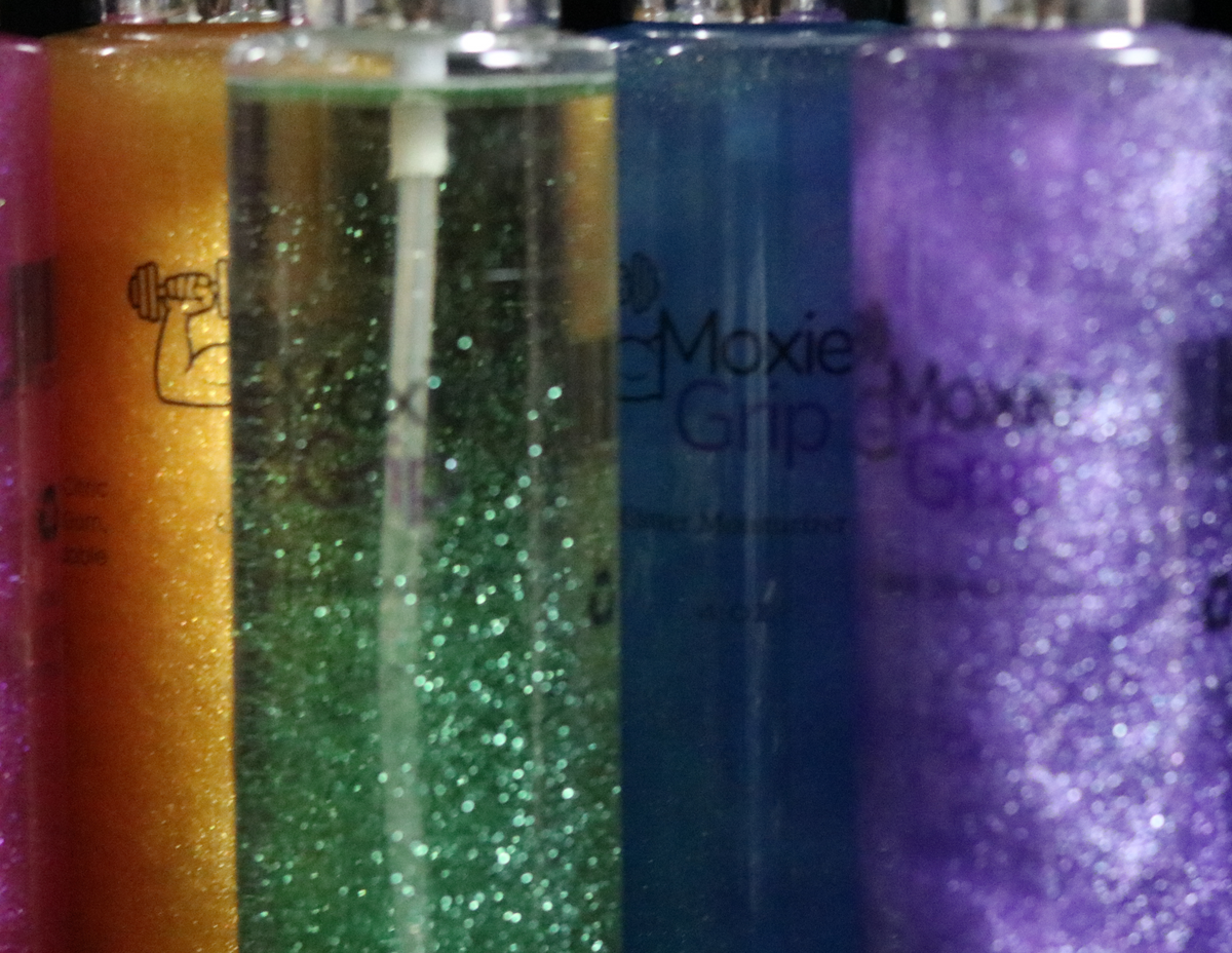 Four glittery grip moisturizer bottles in orange, green, blue, and purple, labeled 'Moxie Grip', shown against a black background.